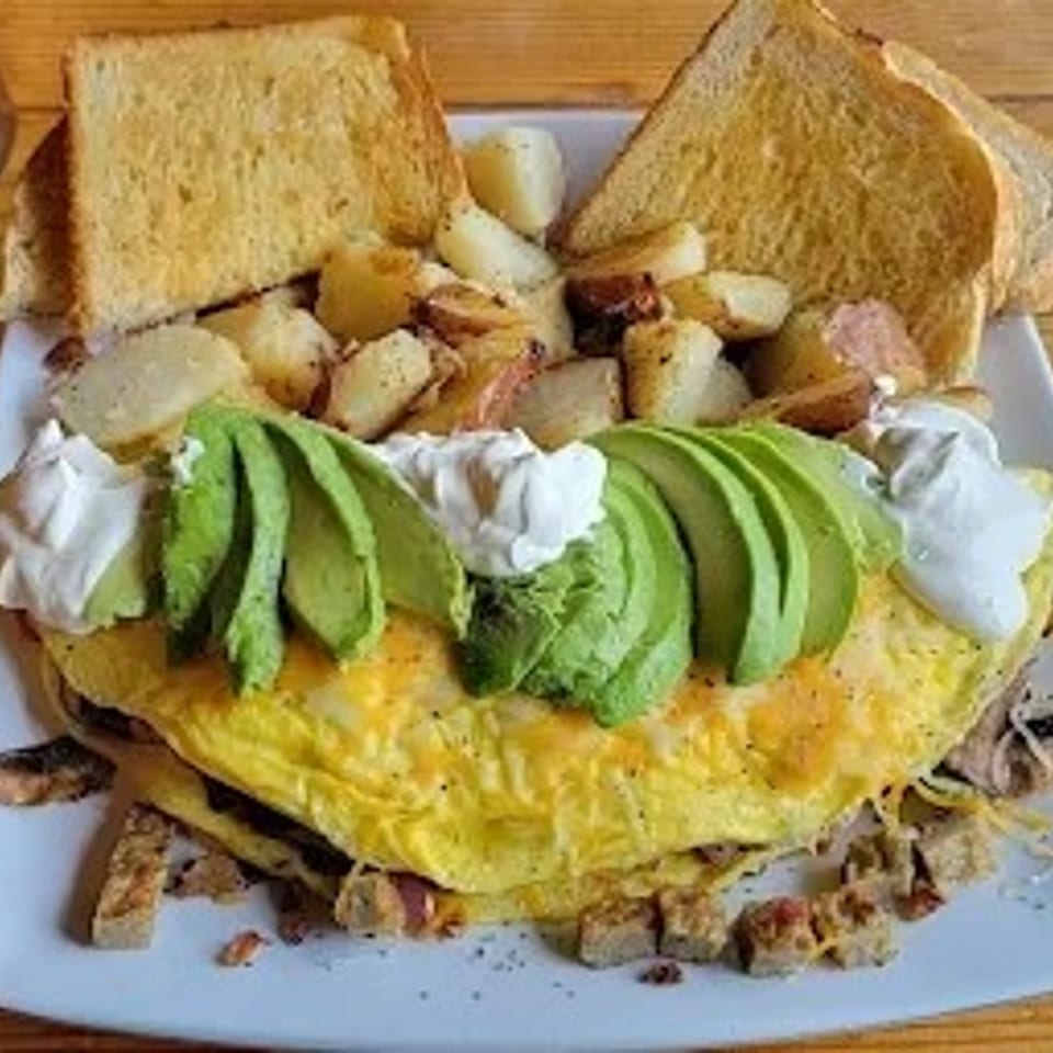 A fluffy omelet topped with avocado slices and sour cream, served with toast and roasted potatoes.