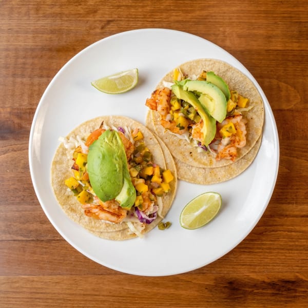 Two shrimp tacos with mango salsa, avocado, and lime wedges on a white plate.