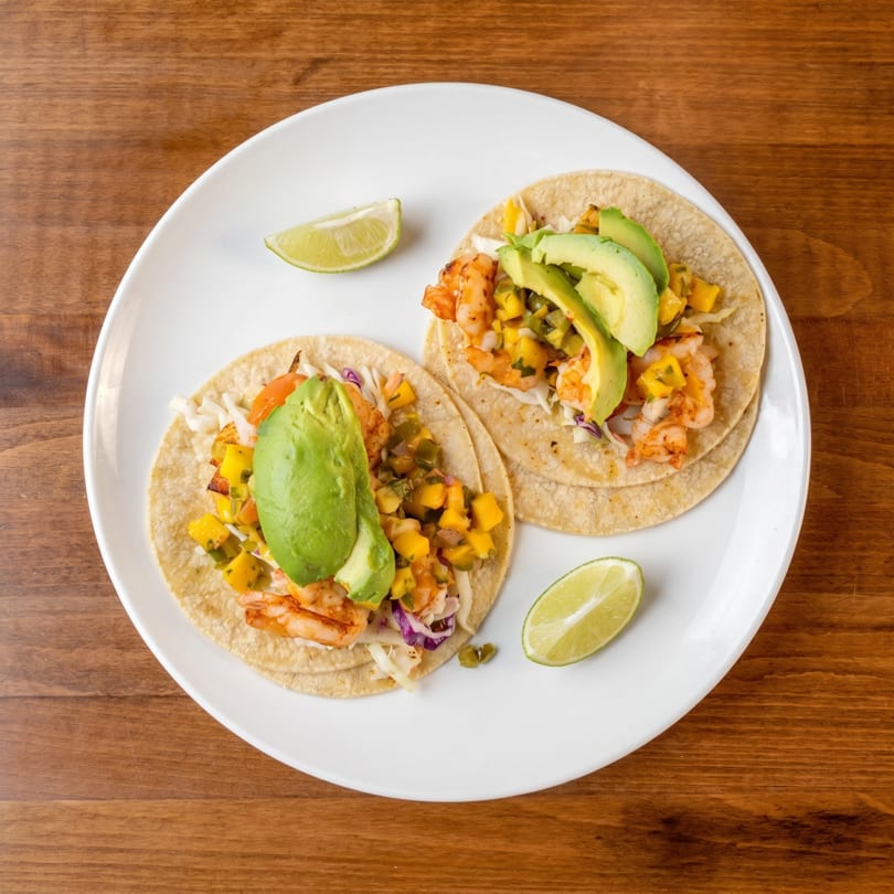 Two shrimp tacos with mango salsa, avocado, and lime wedges on a white plate.
