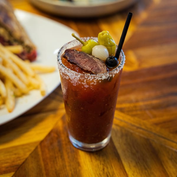 Bloody Mary cocktail garnished with beef jerky, pickled peppers, onions, and olives, next to fries.