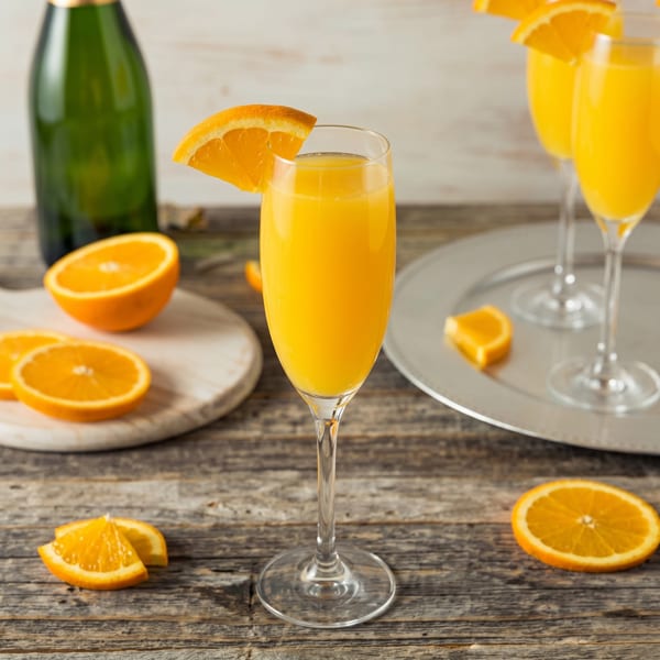 Mimosa cocktails in champagne flutes with orange slices and a green bottle on a wooden table.