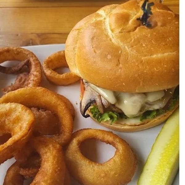 A mushroom swiss burger with lettuce and tomato, served with onion rings and a pickle spear.