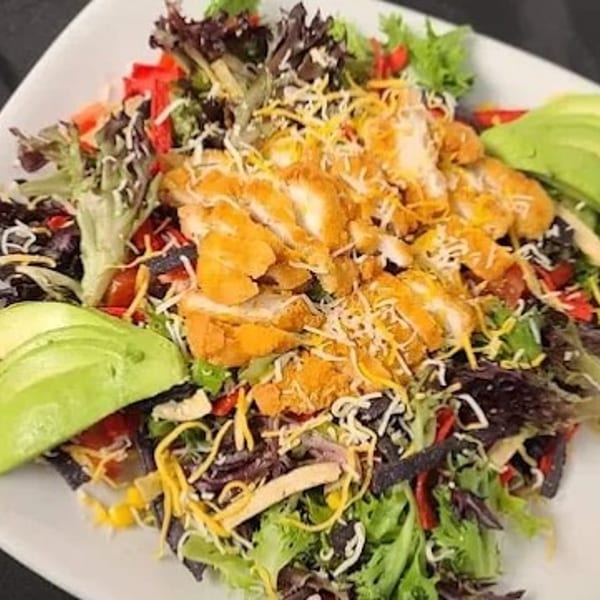 Crispy chicken salad with mixed greens, cheese, avocado, and tortilla strips on a white plate.
