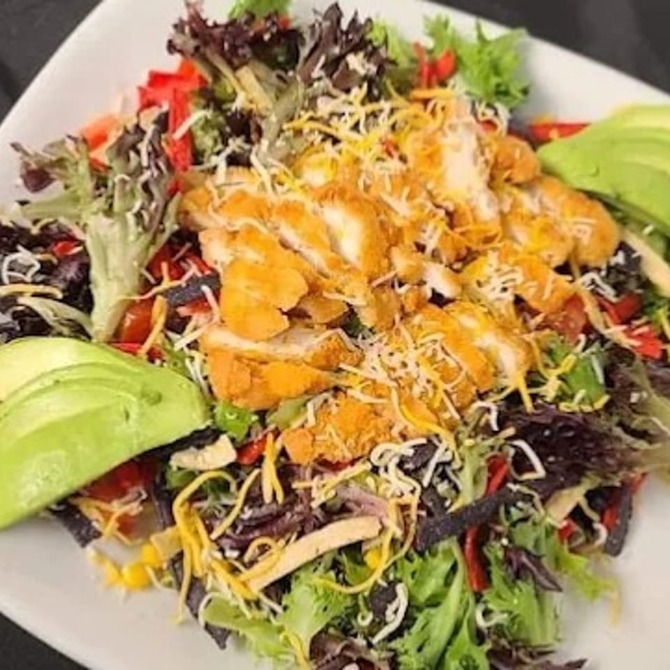 Crispy chicken salad with mixed greens, cheese, avocado, and tortilla strips on a white plate.