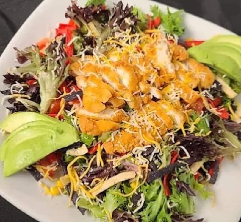 Crispy chicken salad with mixed greens, cheese, avocado, and tortilla strips on a white plate.