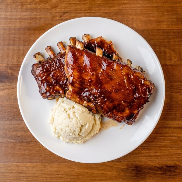 Two racks of BBQ ribs with mashed potatoes on a white plate, on a wooden table.