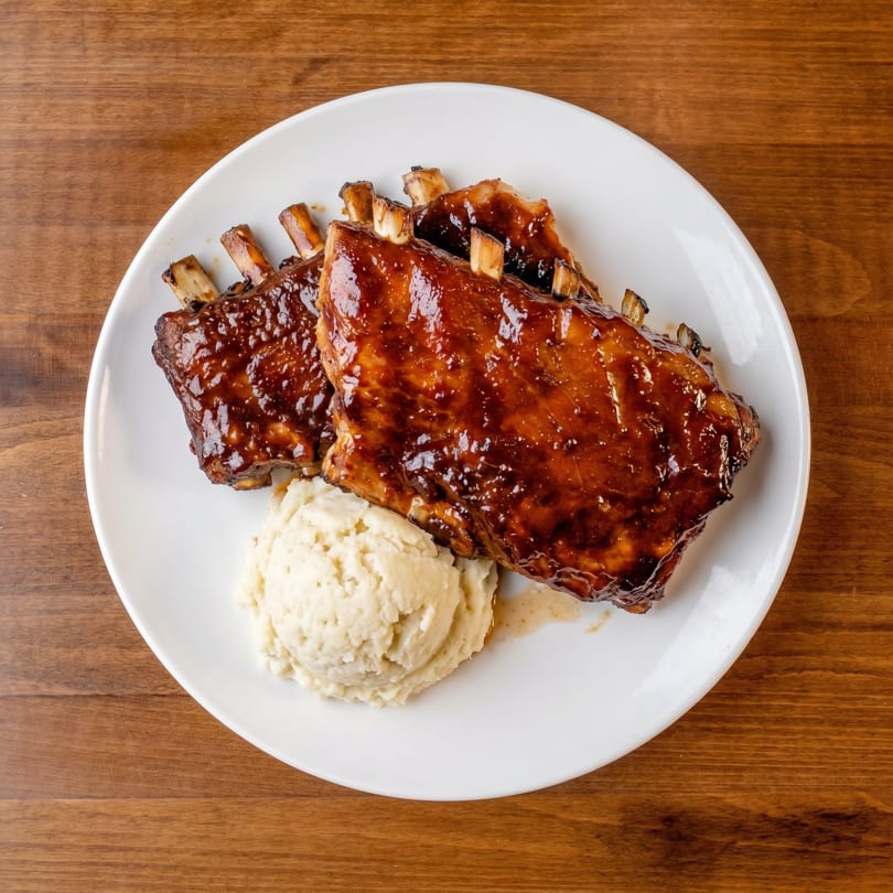 Two racks of BBQ ribs with mashed potatoes on a white plate, on a wooden table.