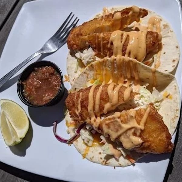 Three crispy fish tacos with slaw, sauce, salsa, and lime on a white plate.