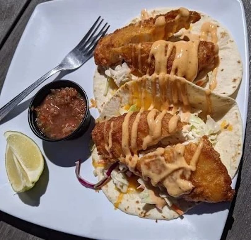 Three crispy fish tacos with slaw, sauce, salsa, and lime on a white plate.