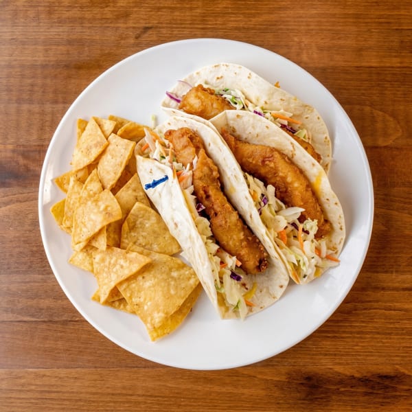 Three fish tacos with coleslaw and a side of tortilla chips on a white plate.