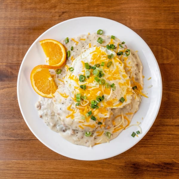 Biscuits and gravy topped with fried eggs, shredded cheese, and green onions, garnished with orange slices.