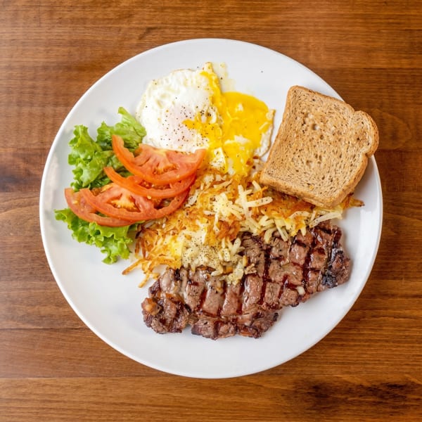 A breakfast plate with grilled steak, hash browns, fried eggs, toast, lettuce, and sliced tomatoes.
