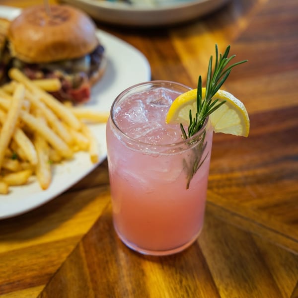 Pink cocktail with lemon and rosemary garnish, next to a plate of sliders and fries.