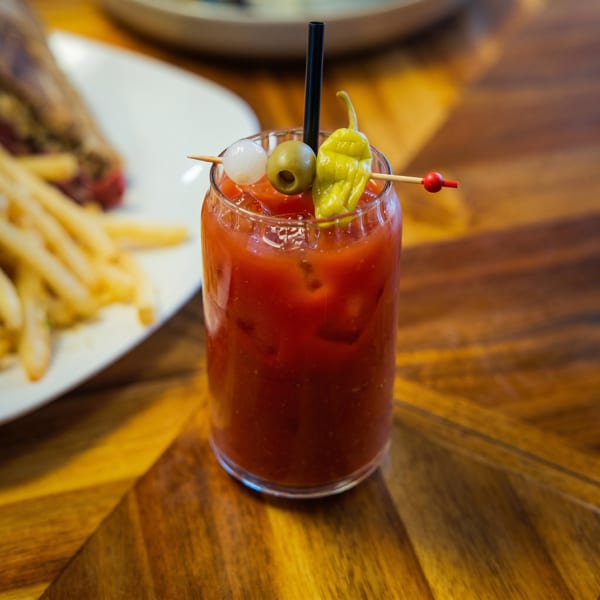 Bloody Mary cocktail garnished with an olive, onion, and pepper, next to a plate of fries.